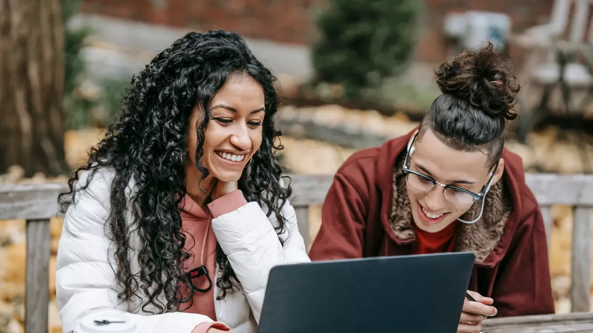 Zwei Studierende sitzen draußen auf einer Bank und schauen gemeinsam auf einen Laptop, umgeben von Pflanzen