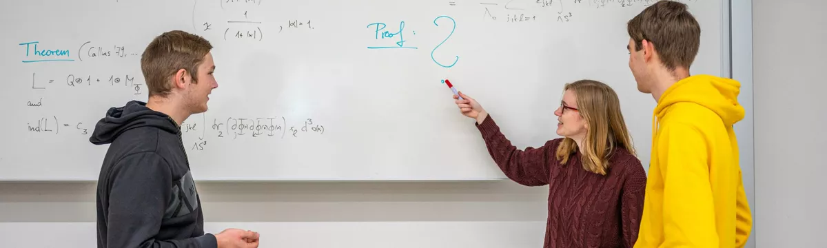 Drei Studierende stehen an einer Tafel und betrachten eine Formel