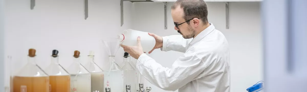 Ein Mann in Schutzkleidung vor mehreren großen Reagenzbehältern, er schüttet eine Flüssigkeit in einen der Behälter
