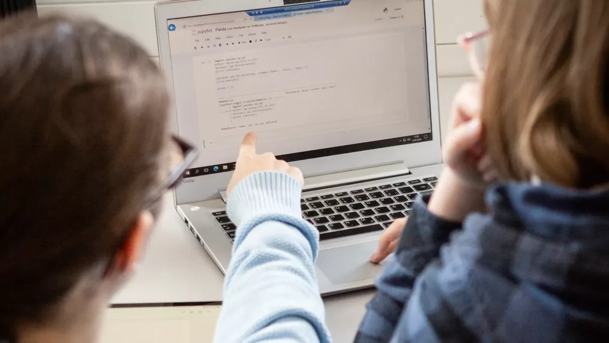Zwei Studierende sitzen vor einem Laptop auf dem HTML-Codes zu sehen sind. Eine Person zeigt auf den Bildschirm.