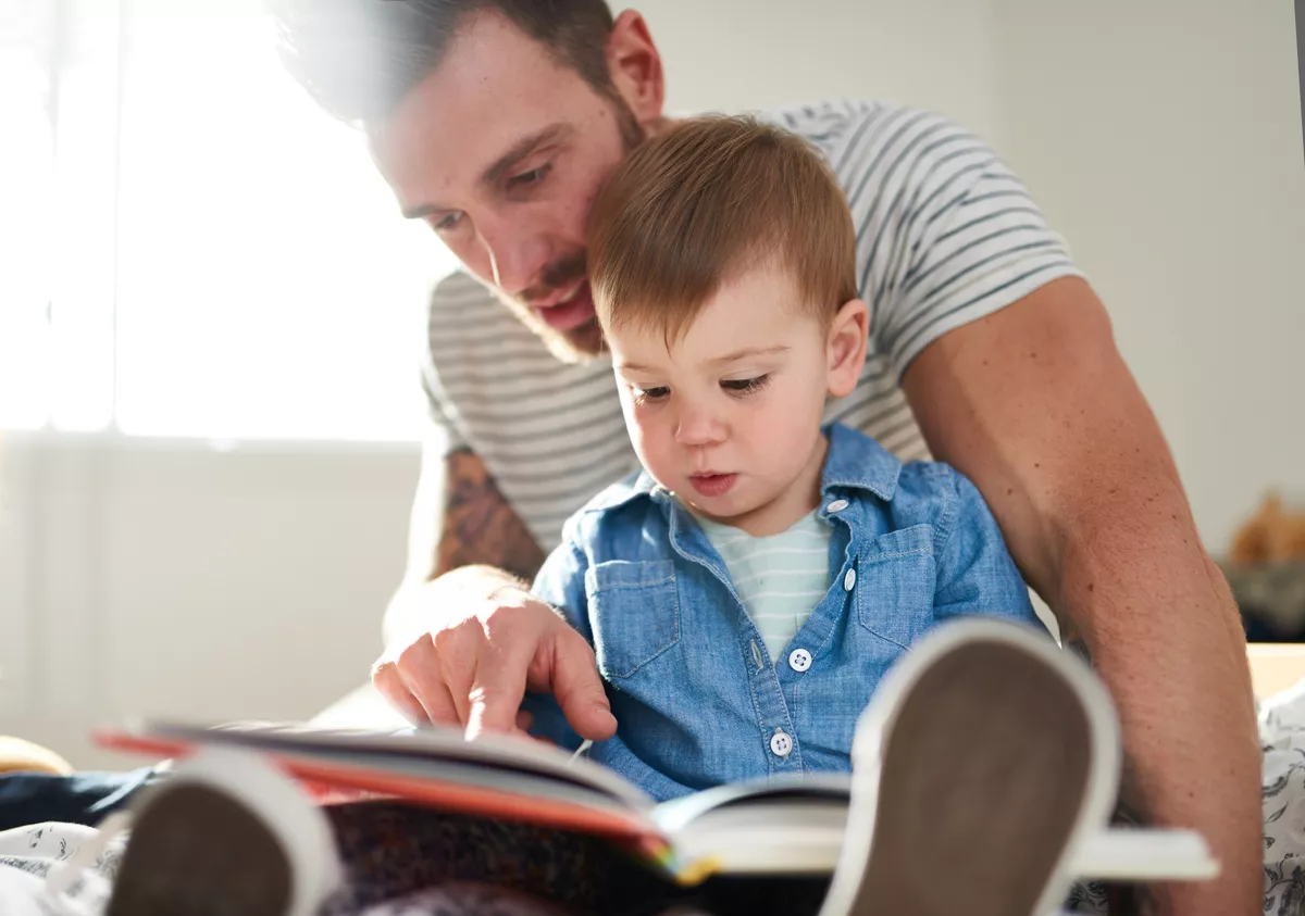 Adorable, young female toddler learning to read book with handsome millennial father on bed