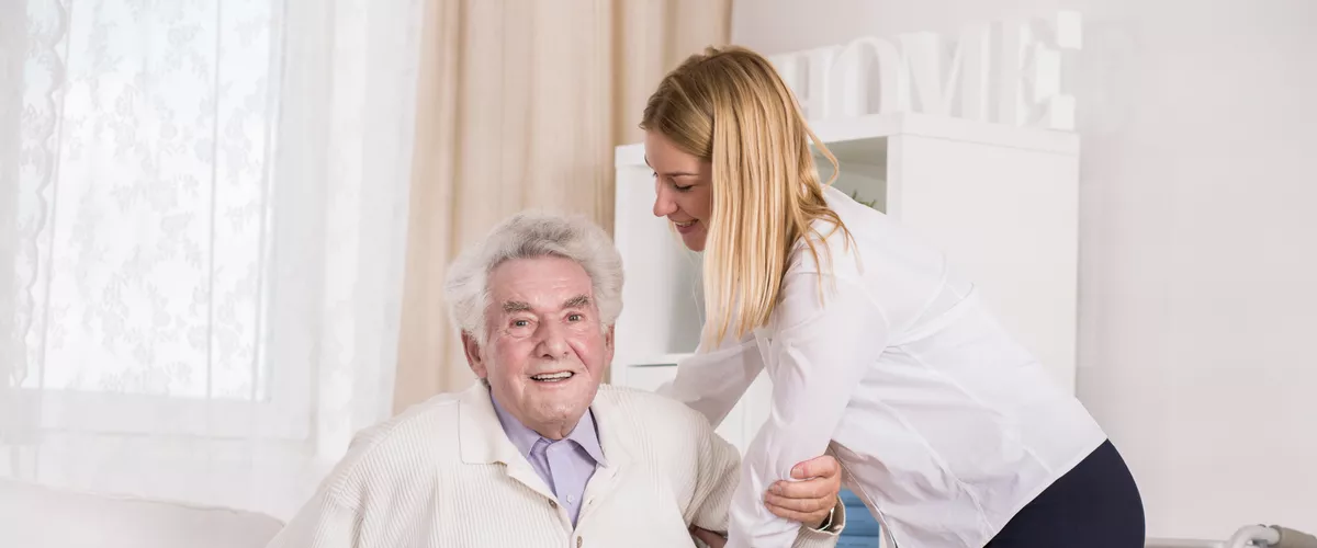 Eine Sozialassistentin hilft einem älteren Herrn beim Aufstehen aus dem Bett.