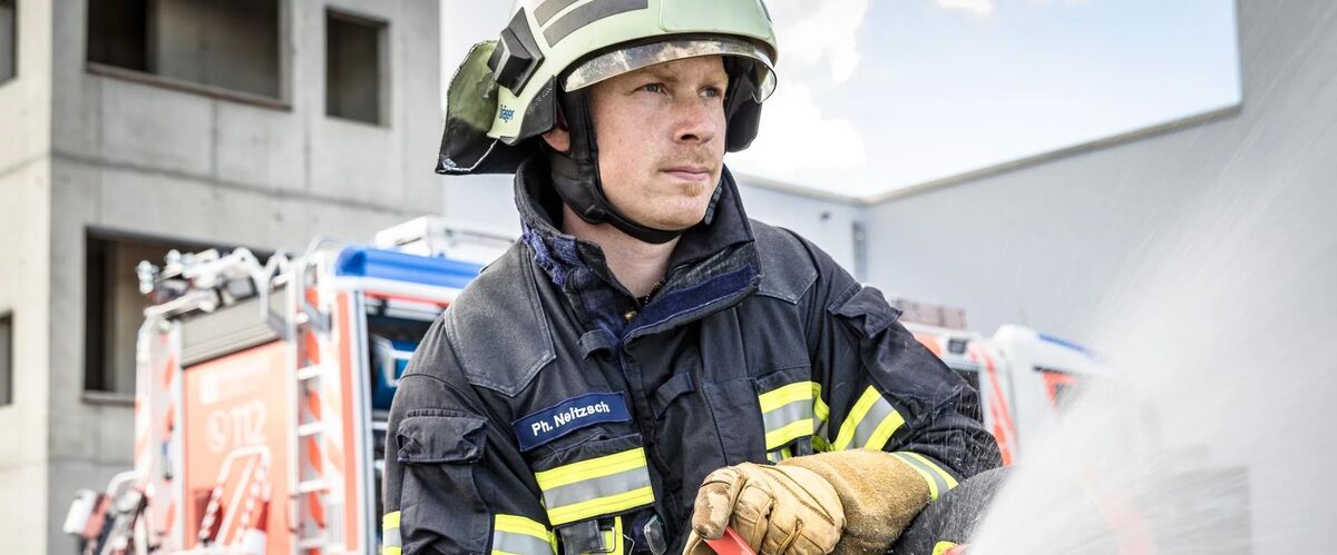 Brandmeister mit Löschschlauch, im Hintergrund Löschfahrzeug